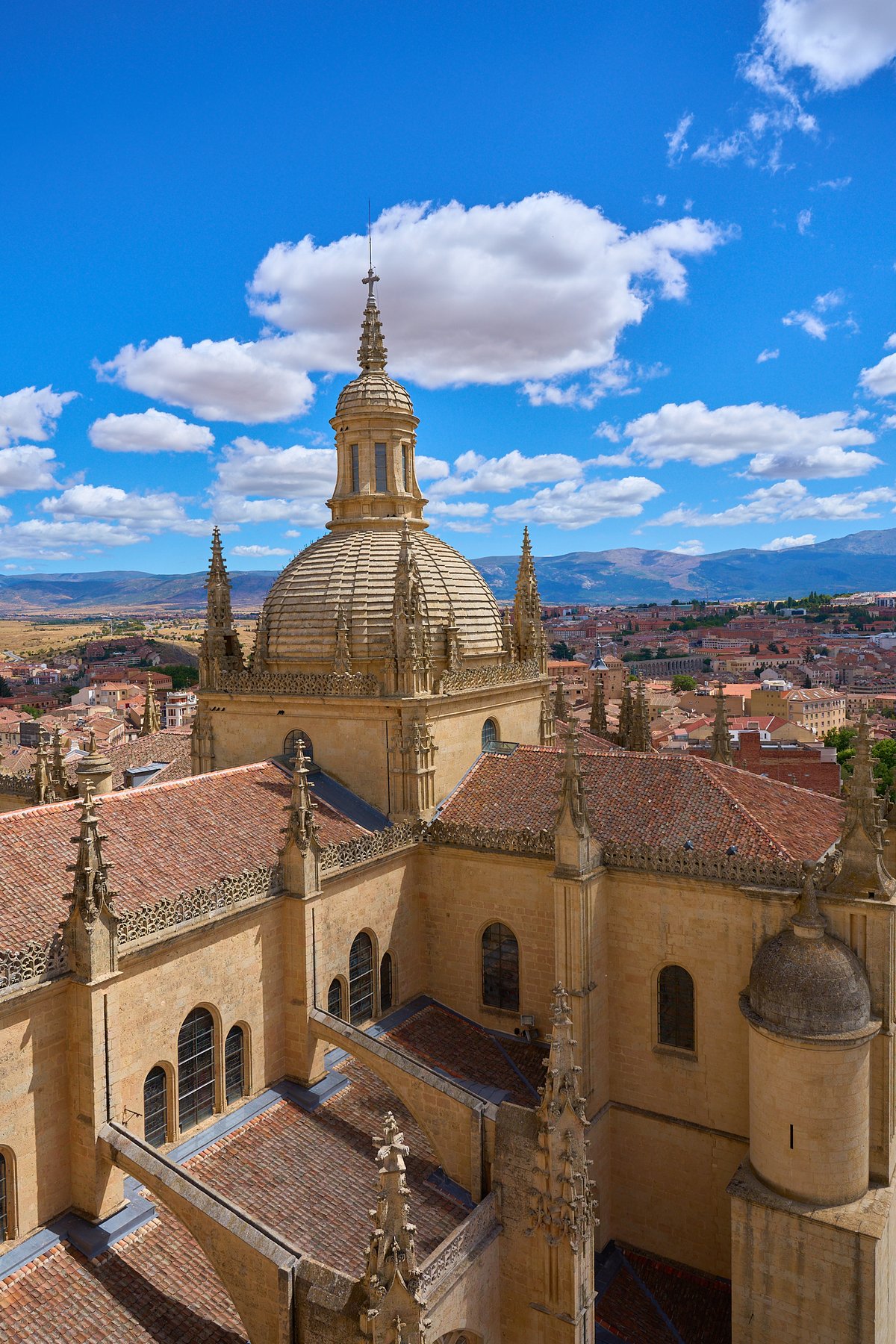 A high-angle view of the ornate stone dome and red-tiled roofs of Segovia Cathedral under a bright blue sky, overlooking the city and distant mountains.