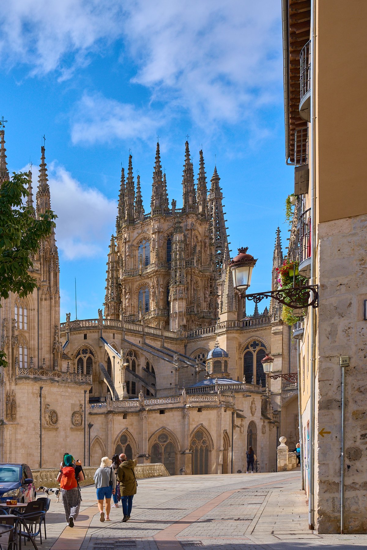Pedestrians walk along a paved street leading toward the intricate Gothic spires of Burgos Cathedral under a bright blue sky.