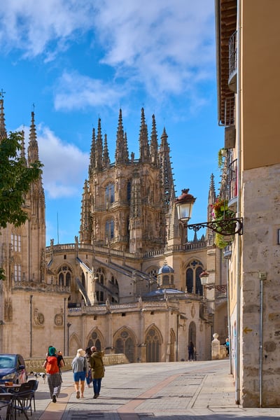 Pedestrians walk along a paved street leading toward the intricate Gothic spires of Burgos Cathedral under a bright blue sky.