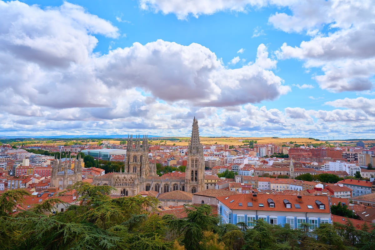 A high-angle view of the Burgos Cathedral and surrounding city with red-tiled roofs under a cloudy blue sky.