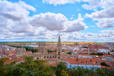A high-angle view of the Burgos Cathedral and surrounding city with red-tiled roofs under a cloudy blue sky.