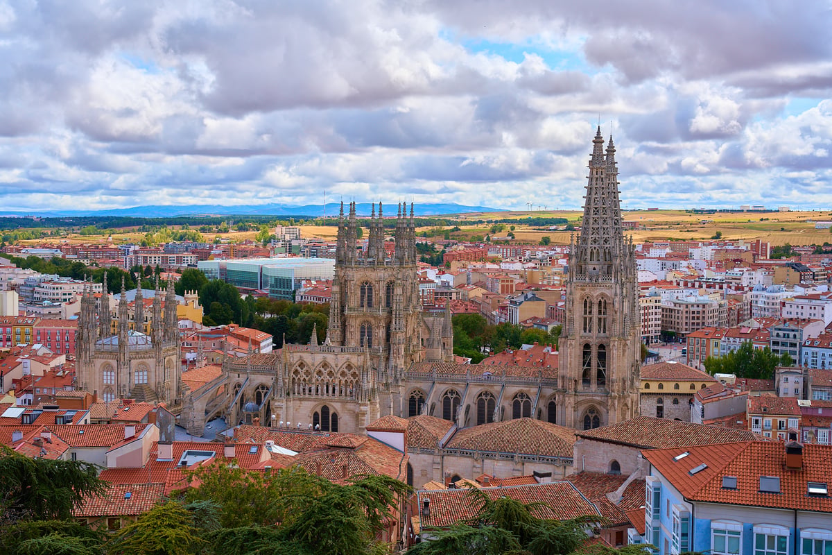A high-angle view of the Gothic Burgos Cathedral towering over a cityscape of red-tiled roofs under a dramatic cloudy sky.