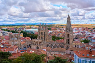 A high-angle view of the Gothic Burgos Cathedral towering over a cityscape of red-tiled roofs under a dramatic cloudy sky.