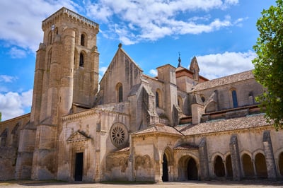 A large Romanesque and Gothic stone monastery with a square bell tower and arched cloisters stands under a bright blue sky with scattered clouds.