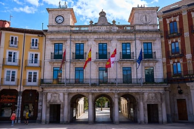 A historic stone town hall with two clock towers and four flags stands above a triple-arched walkway under a blue sky.