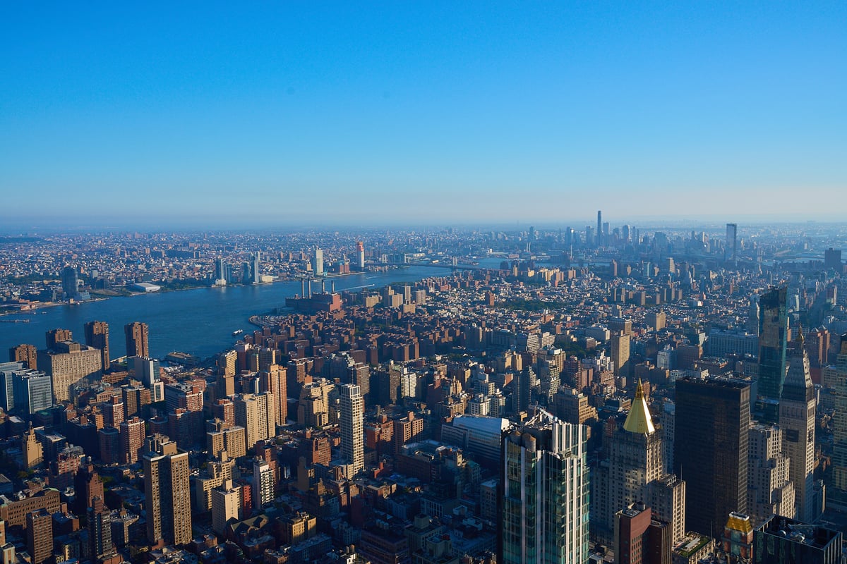An aerial view of the dense Manhattan cityscape overlooking the East River under a clear blue sky in the morning light.