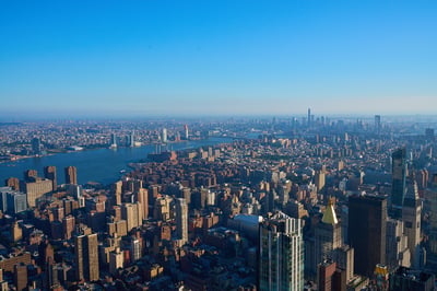 An aerial view of the dense Manhattan cityscape overlooking the East River under a clear blue sky in the morning light.
