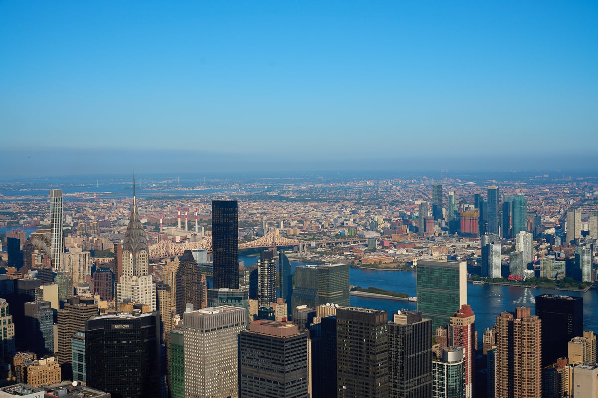 An aerial view of the Midtown Manhattan skyline featuring the Chrysler Building and United Nations Headquarters overlooking the East River under a clear blue sky.