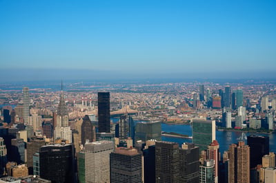 An aerial view of the Midtown Manhattan skyline featuring the Chrysler Building and United Nations Headquarters overlooking the East River under a clear blue sky.