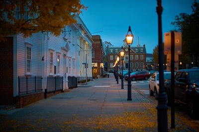 A row of glowing street lamps lines a sidewalk in a Newport, RI at dusk, leading toward a brick building with a clock tower.