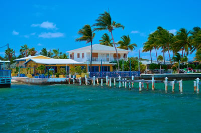 A yellow waterfront building and white two-story house stand among palm trees beside a turquoise sea with a wooden pier in the foreground.