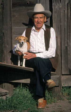 Tom Dorrance seated with his dog outside a ranch barn