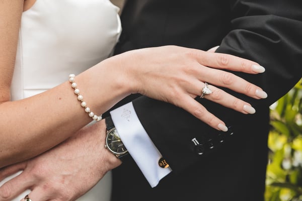 Close-up of the bride's hand resting on the groom's arm, showing her pear-cut diamond engagement ring, pearl bracelet, and his watch and cufflinks