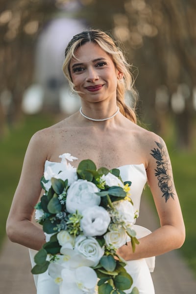 Close-up three-quarter portrait of bride on the tree-lined path holding her bouquet, smiling directly at camera in warm light