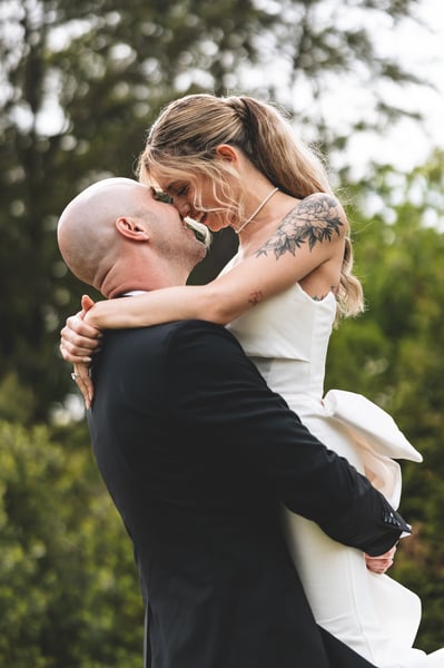 Groom lifting and embracing the bride, their foreheads nearly touching as they share a joyful moment outdoors against green trees