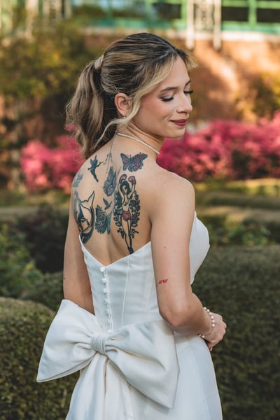 Bride turned away in the formal garden, showing her colorful back tattoos and large gown bow, pink azaleas brilliant in the background