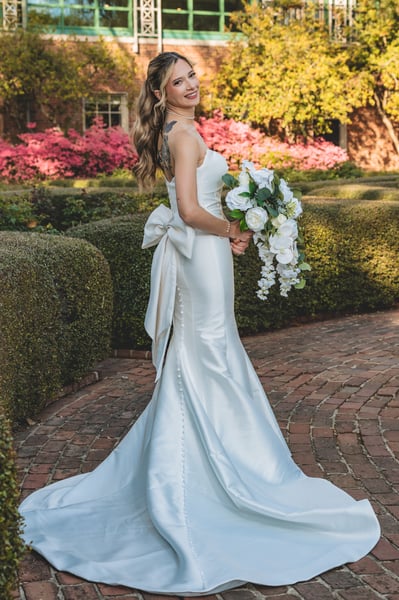 Bride turned away on a formal garden brick path, large gown bow and colorful back tattoos visible, bouquet held to the side, azaleas behind