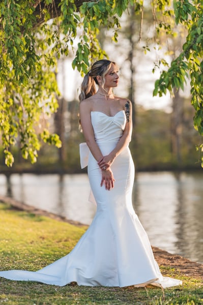 Bride standing under willow branches at the lakeside in golden hour, three-quarter view with hands clasped