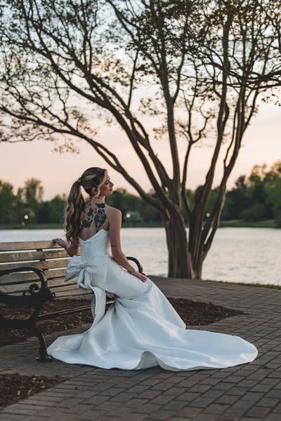 Bride seated on a park bench at dusk, gown train spread out behind her, back tattoos showing above the strapless neckline, lake behind
