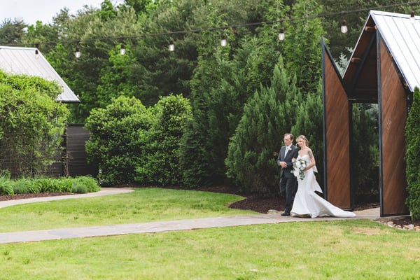 Bride being walked through the venue grounds by a man in a dark suit, string lights and the wedding pavilion visible in the lush background