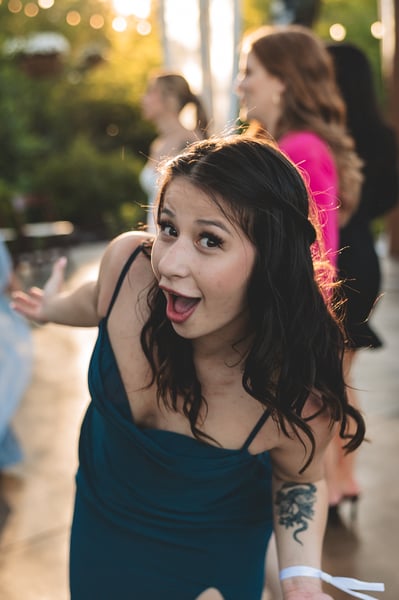 A guest in a teal dress making a wide-eyed, exuberant expression at the camera during the reception, golden bokeh string lights behind her
