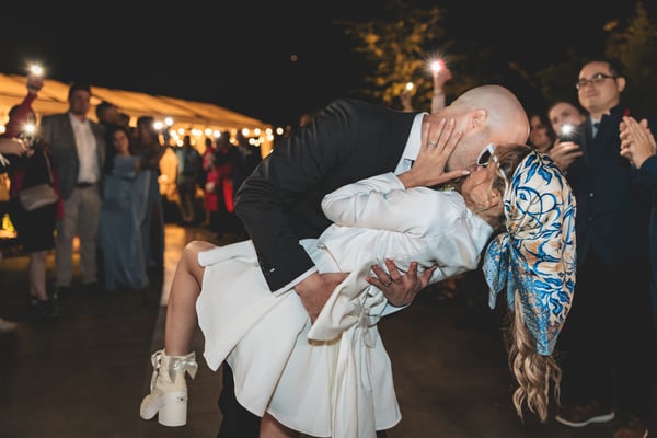 Groom dipping the bride for a dramatic kiss during the sparkler send-off, guests surrounding them with lit sparklers in the night air, the bride in her short white dress and heart sunglasses