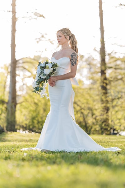 Bride in mermaid gown standing in an open park field with bouquet, backlit by bright morning sun