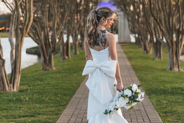Bride on the brick tree-lined path at dusk, back to camera, large bow and tattoos visible, bouquet in her lowered hand