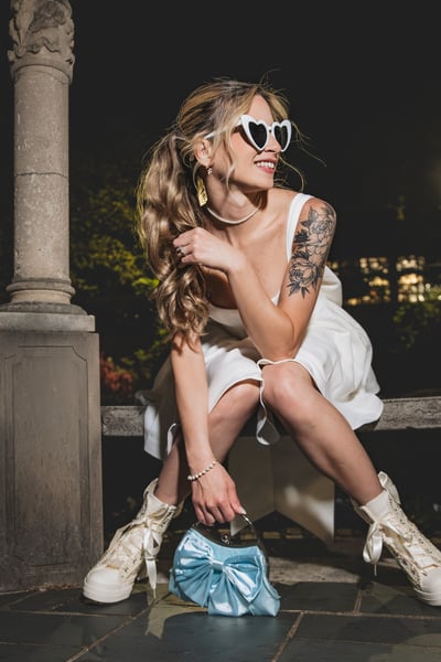 Bride in short white dress laughing and seated on stone steps in the gazebo at night, heart sunglasses on, blue bow bag and bejeweled Converse