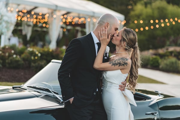 Bride standing through the open top of a vintage Corvette while the groom drives, both poised and elegant at the venue entrance at dusk