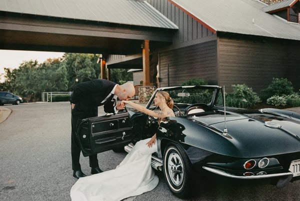 Groom bending to kiss the brides hand as she sits in the open Corvette, the venues covered drive arching behind them