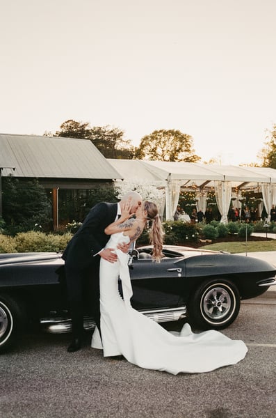 Bride and groom kissing beside the Corvette with the stone chimney and lit reception tent visible behind them