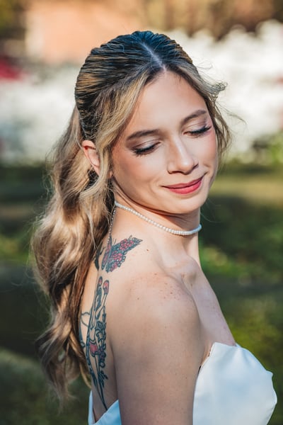 Close-up portrait of bride from slightly behind, eyes closed peacefully, pearl choker visible, colorful back tattoo partially in frame