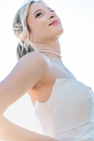 Close-up portrait of bride looking up and smiling broadly, bright overexposed sky behind her, pearl choker catching the light