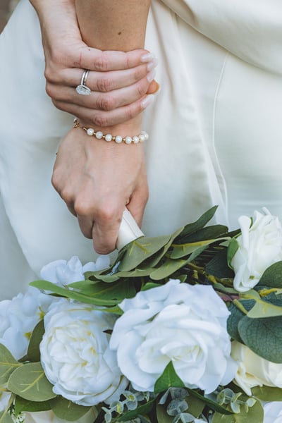 Close-up of bride's hands holding the stem of her bouquet, engagement ring and pearl bracelet visible against the white gown