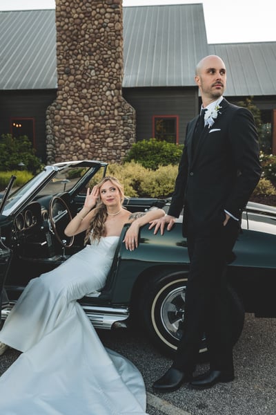 Bride seated in a vintage dark green Corvette convertible beside the standing groom, with a stone building in the background at dusk