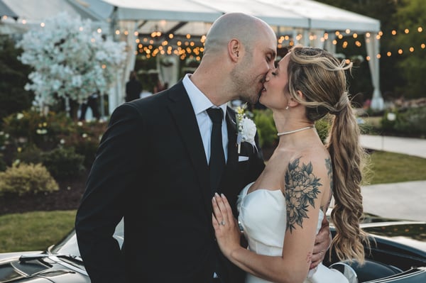 Bride and groom kissing in front of the vintage Corvette at dusk with reception string lights glowing warmly in the background
