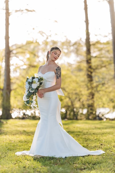 Bride in strapless mermaid gown standing in a sun-drenched park, eyes downcast, holding bouquet, trees in the background