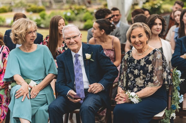Three smiling guests — two women and an older man wearing a boutonniere — seated in the front row at the outdoor ceremony