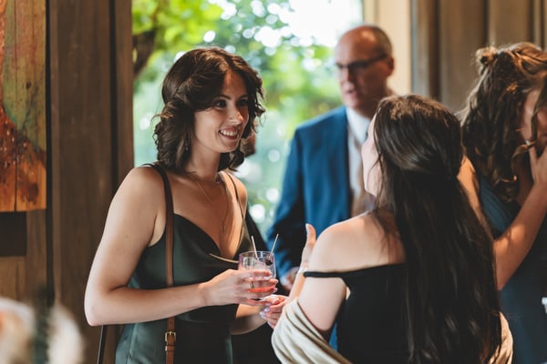 Two female guests in elegant dark dresses chatting and smiling during cocktail hour, one holding a cocktail