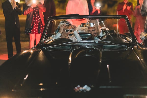 Side view of the vintage dark green Corvette in motion at night, a large white bow on the trunk and the bride visible in the passenger seat