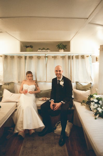 Bride and groom seated inside a vintage Airstream trailer with striped curtains and plants overhead