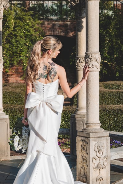 Bride turned away inside the ornate garden gazebo, large back bow and colorful back tattoos showing, reaching for a stone column