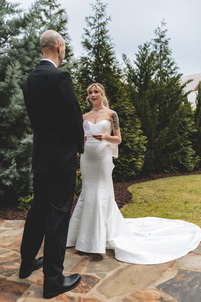Bride smiling warmly while reading her vows to the groom on a stone patio surrounded by tall green evergreens