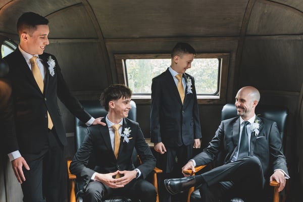 Groom and three younger groomsmen in dark suits with gold ties and boutonnieres, laughing together inside a vintage aircraft interior