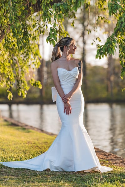 Bride standing under drooping willow branches at the lakeside, hands clasped in front, looking to the side in warm evening light