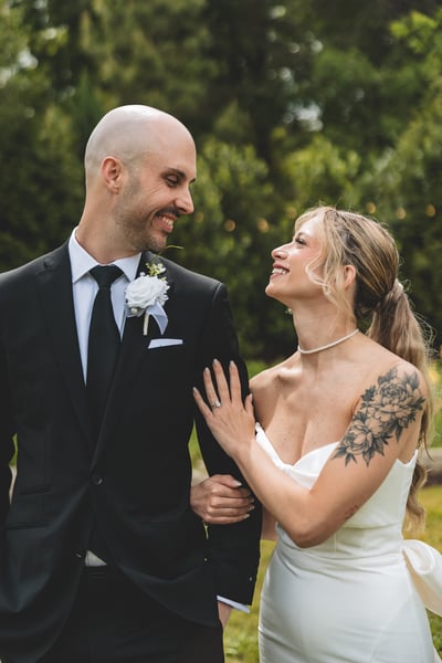 Bride and groom gazing at each other and laughing softly in a lush garden setting, she in her white gown and he in his dark suit with boutonniere