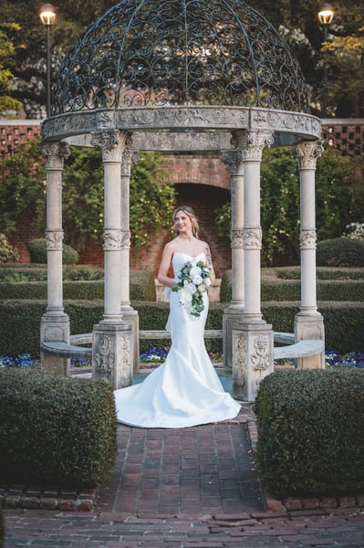 Full-length portrait of bride inside the iron-dome garden gazebo, bouquet held forward, blue pansies and boxwood visible through the columns