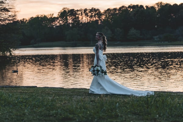 Wide silhouette shot of bride walking along the lake's edge at sunset carrying her bouquet, dramatic orange and pink sky reflected in the water