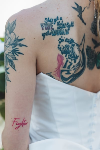 Close-up of bride's back tattoos including a mermaid, the word "Fighter" in red, and the phrase "She's got a fire that could light up the world"